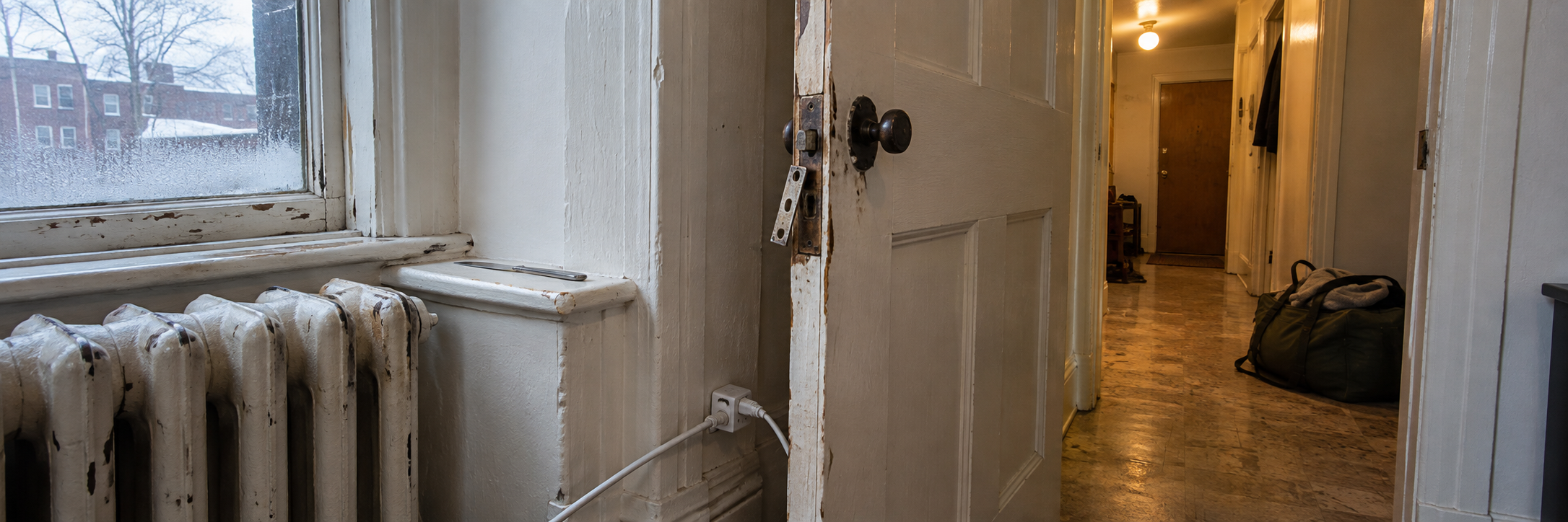 A worn radiator and half-open door lead from cold window light into a warm hallway with an overnight bag.